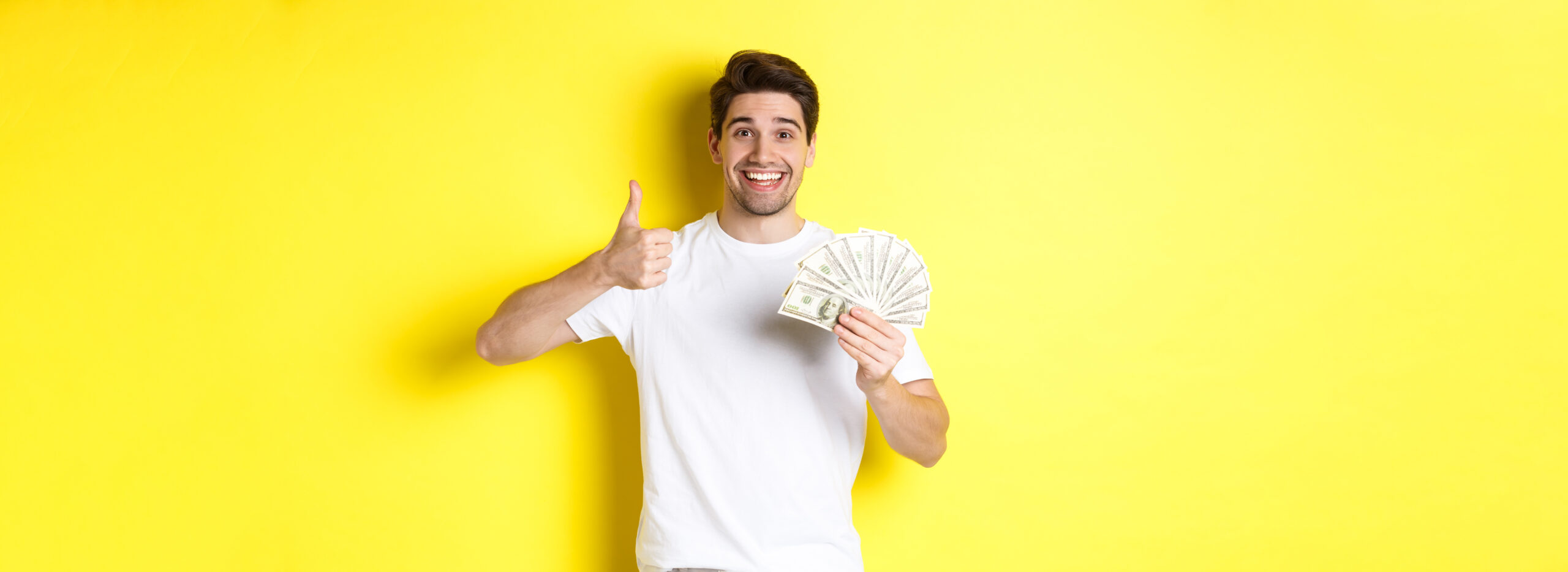Satisfied buyer man showing thumb-up and holding money, shopping with cash, standing over yellow background.