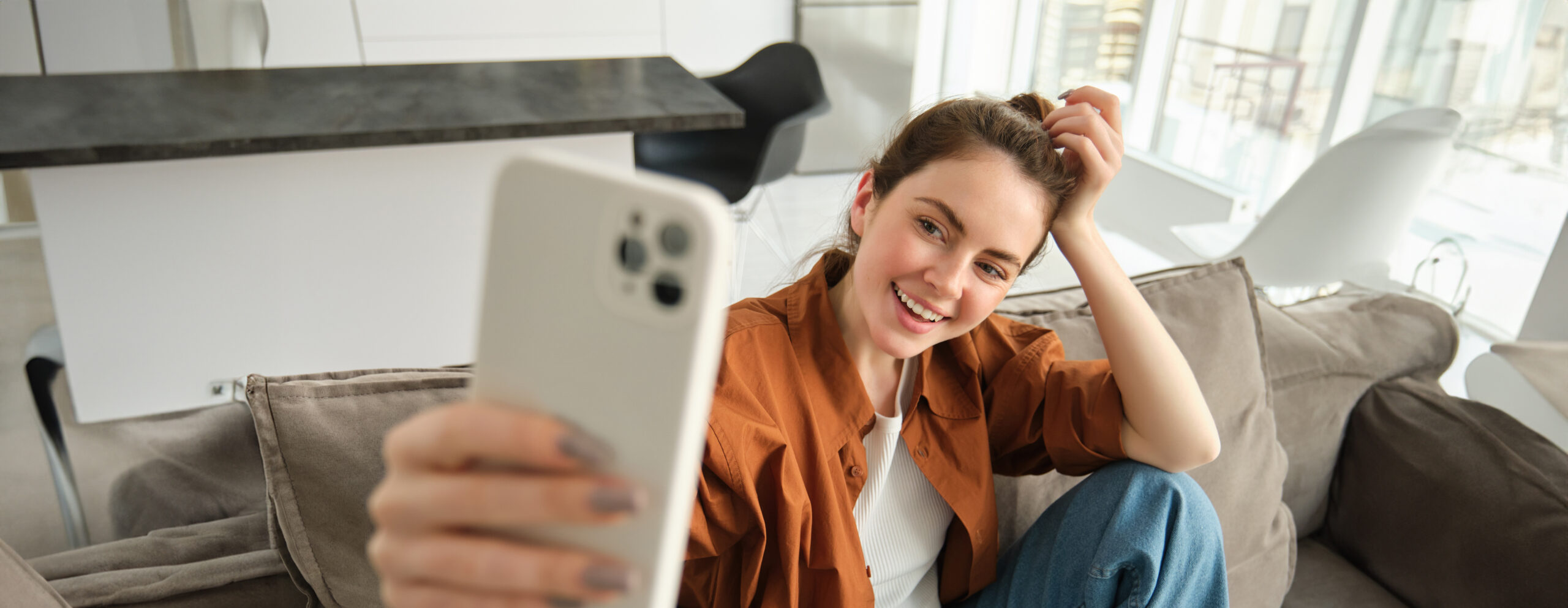 Portrait of beautiful, happy young female model, sitting on couch with smartphone, taking selfie on mobile phone, making cheerful smiling face.